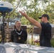 Two men are engaged in a discussion near a marine radar dome unit, likely branded 'Furuno', mounted on a mast. One man wearing a black uniform with rank insignia and a lanyard listens attentively while the other, in a black t-shirt and cap, gestures expressively with one hand. The setting appears to be an outdoor area near a marina or technical facility.