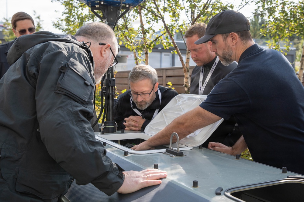 Four men are gathered around what appears to be a piece of technical or military equipment, examining an open hatch or access panel on its surface. The setting is outdoors with trees and modular buildings visible in the background. The individuals appear to be technicians or specialists conducting an inspection or demonstration.