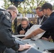 Four men are gathered around what appears to be a piece of technical or military equipment, examining an open hatch or access panel on its surface. The setting is outdoors with trees and modular buildings visible in the background. The individuals appear to be technicians or specialists conducting an inspection or demonstration.