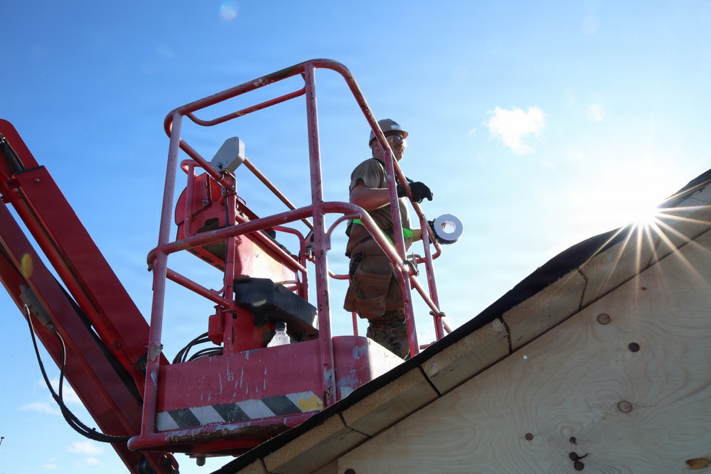 A construction worker stands in the basket of a red aerial lift (boom lift) at elevation, working near a roofline. The worker is wearing a white hard hat and appears to be holding a roll of tape or similar material. The photo is taken from a low angle looking upward, with bright sunlight creating a starburst effect in the upper right.