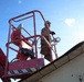 A construction worker stands in the basket of a red aerial lift (boom lift) at elevation, working near a roofline. The worker is wearing a white hard hat and appears to be holding a roll of tape or similar material. The photo is taken from a low angle looking upward, with bright sunlight creating a starburst effect in the upper right.
