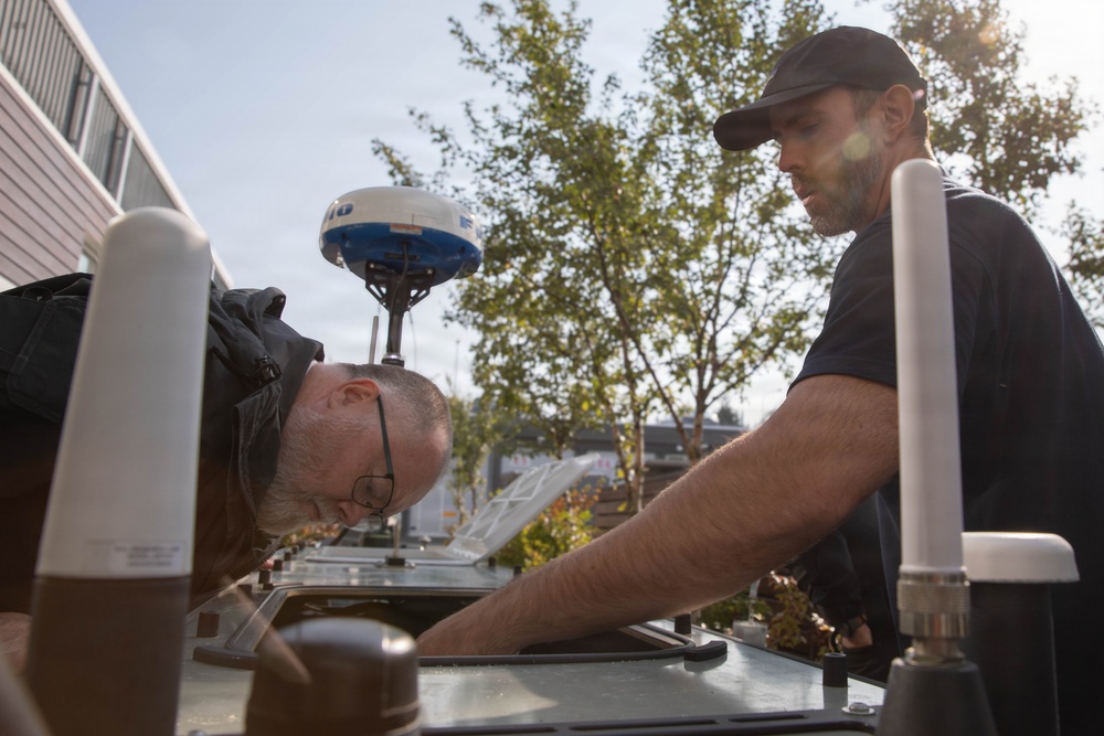 Two men are working together on what appears to be the roof or top surface of a vehicle or vessel equipped with navigation/communication equipment. One man is leaning down closely inspecting or working on a surface while the other assists from the opposite side. The setting appears to be an outdoor marina or industrial area with trees and buildings visible in the background.