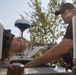 Two men are working together on what appears to be the roof or top surface of a vehicle or vessel equipped with navigation/communication equipment. One man is leaning down closely inspecting or working on a surface while the other assists from the opposite side. The setting appears to be an outdoor marina or industrial area with trees and buildings visible in the background.