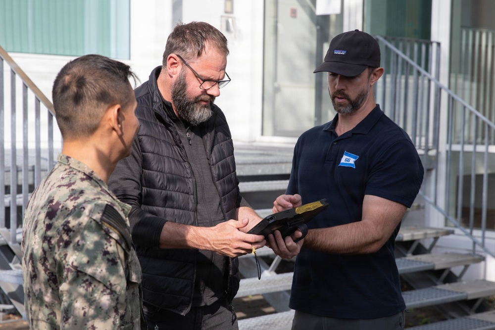 Three individuals are gathered outdoors near a building with metal staircases, examining a small device or object together. One person is wearing a military camouflage uniform, while the other two are in civilian attire. The interaction appears to be a briefing or demonstration involving a handheld item.