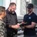 Three individuals are gathered outdoors near a building with metal staircases, examining a small device or object together. One person is wearing a military camouflage uniform, while the other two are in civilian attire. The interaction appears to be a briefing or demonstration involving a handheld item.