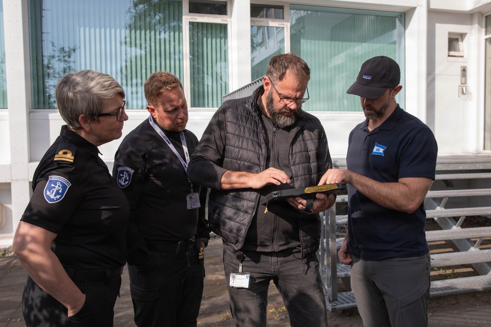 Four individuals are gathered outdoors in front of a white building, examining a tablet or clipboard device together. Two of the individuals are wearing black uniforms with anchor insignia patches, suggesting maritime or coast guard affiliation, while the other two are in civilian attire. The group appears to be reviewing documents or data on the device in a professional context.