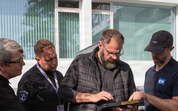 Four individuals are gathered outdoors in front of a white building, examining a tablet or clipboard device together. Two of the individuals are wearing black uniforms with anchor insignia patches, suggesting maritime or coast guard affiliation, while the other two are in civilian attire. The group appears to be reviewing documents or data on the device in a professional context.