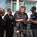 Four individuals are gathered outdoors in front of a white building, examining a tablet or clipboard device together. Two of the individuals are wearing black uniforms with anchor insignia patches, suggesting maritime or coast guard affiliation, while the other two are in civilian attire. The group appears to be reviewing documents or data on the device in a professional context.