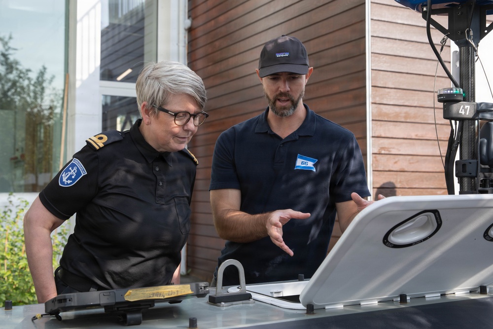 Two individuals are examining equipment on what appears to be a vessel or maritime platform outdoors. One person is wearing a dark uniform with an anchor insignia patch, suggesting a maritime authority or coast guard role, while the other wears a dark polo shirt with a logo badge numbered '66' and a Patagonia cap. The man appears to be explaining or demonstrating something about the equipment to the uniformed individual.