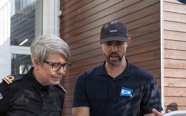 Two individuals are examining equipment on what appears to be a vessel or maritime platform outdoors. One person is wearing a dark uniform with an anchor insignia patch, suggesting a maritime authority or coast guard role, while the other wears a dark polo shirt with a logo badge numbered '66' and a Patagonia cap. The man appears to be explaining or demonstrating something about the equipment to the uniformed individual.