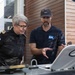Two individuals are examining equipment on what appears to be a vessel or maritime platform outdoors. One person is wearing a dark uniform with an anchor insignia patch, suggesting a maritime authority or coast guard role, while the other wears a dark polo shirt with a logo badge numbered '66' and a Patagonia cap. The man appears to be explaining or demonstrating something about the equipment to the uniformed individual.