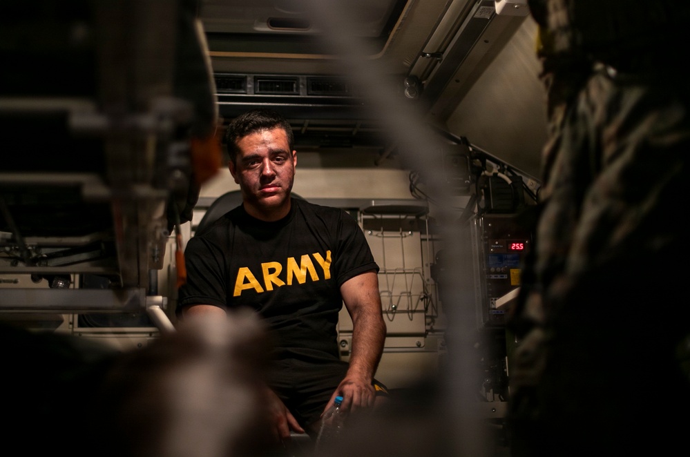 A man wearing a black U.S. Army physical training shirt is seated inside what appears to be a military vehicle or enclosed equipment bay. He has visible marks or bruising on his face and appears to be in a confined, equipment-filled space. The image is shot through a narrow gap, creating a framed, documentary-style composition.