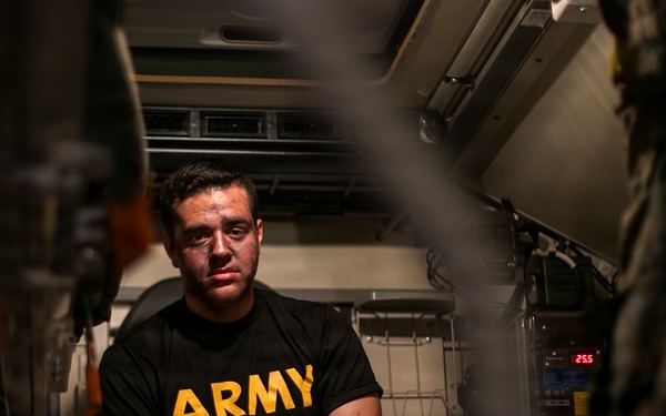 A man wearing a black U.S. Army physical training shirt is seated inside what appears to be a military vehicle or enclosed equipment bay. He has visible marks or bruising on his face and appears to be in a confined, equipment-filled space. The image is shot through a narrow gap, creating a framed, documentary-style composition.