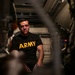 A man wearing a black U.S. Army physical training shirt is seated inside what appears to be a military vehicle or enclosed equipment bay. He has visible marks or bruising on his face and appears to be in a confined, equipment-filled space. The image is shot through a narrow gap, creating a framed, documentary-style composition.