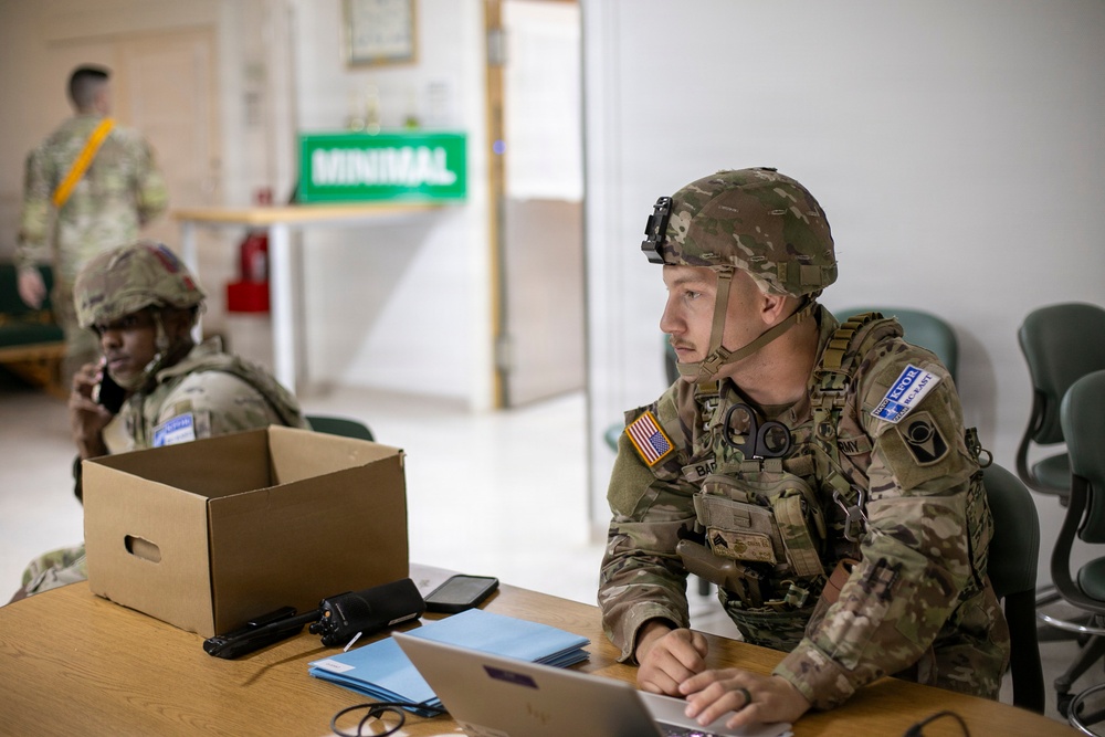 Two U.S. Army soldiers in full combat gear, including helmets and tactical vests, are seated at a wooden table in what appears to be a military operations or command post environment. The foreground soldier is working on a laptop computer while wearing an American flag patch and 'ARMY' tape on his uniform. A third soldier is visible in the background near a green sign reading 'MINIMAL'.