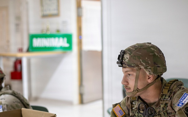 Two U.S. Army soldiers in full combat gear, including helmets and tactical vests, are seated at a wooden table in what appears to be a military operations or command post environment. The foreground soldier is working on a laptop computer while wearing an American flag patch and 'ARMY' tape on his uniform. A third soldier is visible in the background near a green sign reading 'MINIMAL'.