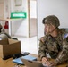 Two U.S. Army soldiers in full combat gear, including helmets and tactical vests, are seated at a wooden table in what appears to be a military operations or command post environment. The foreground soldier is working on a laptop computer while wearing an American flag patch and 'ARMY' tape on his uniform. A third soldier is visible in the background near a green sign reading 'MINIMAL'.