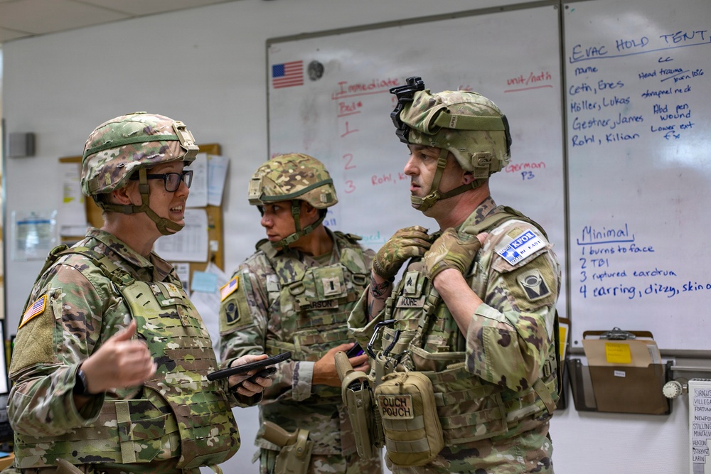 Three U.S. Army soldiers in full combat gear, including helmets and body armor, are gathered in what appears to be a military operations or medical briefing room. They are engaged in a discussion, with one soldier holding a tablet device. A whiteboard behind them displays medical triage information including 'EVAC HOLD TENT' categories and casualty names with injuries.