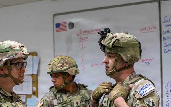 Three U.S. Army soldiers in full combat gear, including helmets and body armor, are gathered in what appears to be a military operations or medical briefing room. They are engaged in a discussion, with one soldier holding a tablet device. A whiteboard behind them displays medical triage information including 'EVAC HOLD TENT' categories and casualty names with injuries.