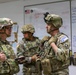 Three U.S. Army soldiers in full combat gear, including helmets and body armor, are gathered in what appears to be a military operations or medical briefing room. They are engaged in a discussion, with one soldier holding a tablet device. A whiteboard behind them displays medical triage information including 'EVAC HOLD TENT' categories and casualty names with injuries.