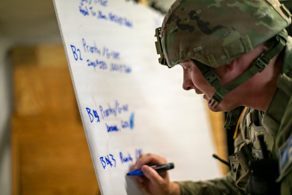 A soldier in full combat gear, including a camouflage helmet and tactical vest, is writing on a large whiteboard or flip chart with a blue marker. The whiteboard contains handwritten notes organized into sections labeled B2, B1g, and B43, appearing to reference priorities or orders. This appears to be a military planning or briefing session.
