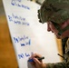 A soldier in full combat gear, including a camouflage helmet and tactical vest, is writing on a large whiteboard or flip chart with a blue marker. The whiteboard contains handwritten notes organized into sections labeled B2, B1g, and B43, appearing to reference priorities or orders. This appears to be a military planning or briefing session.