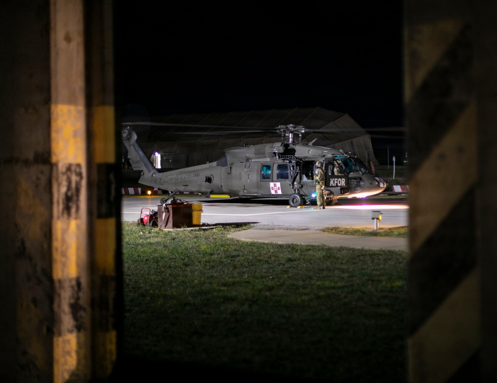 A United States Army helicopter, marked with 'KFOR' (Kosovo Force) and a medical red cross symbol, is parked on a tarmac at night. The image is captured through a gap in what appears to be a concrete or wooden barrier with yellow safety markings. At least one soldier in full combat gear is visible near the aircraft.