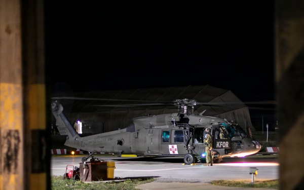 A United States Army helicopter, marked with 'KFOR' (Kosovo Force) and a medical red cross symbol, is parked on a tarmac at night. The image is captured through a gap in what appears to be a concrete or wooden barrier with yellow safety markings. At least one soldier in full combat gear is visible near the aircraft.