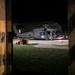 A United States Army helicopter, marked with 'KFOR' (Kosovo Force) and a medical red cross symbol, is parked on a tarmac at night. The image is captured through a gap in what appears to be a concrete or wooden barrier with yellow safety markings. At least one soldier in full combat gear is visible near the aircraft.
