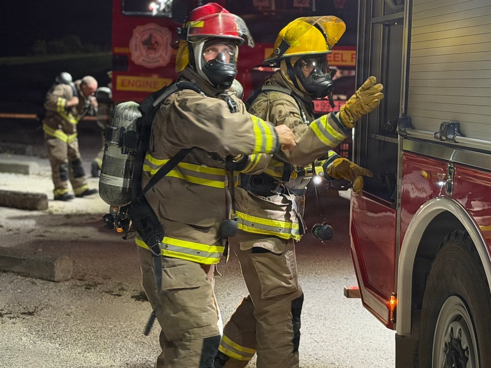 A nighttime firefighting scene showing two fully equipped firefighters in the foreground wearing SCBA (Self-Contained Breathing Apparatus) gear, turnout suits, and full-face respirator masks. One firefighter wears a red helmet and the other a yellow helmet, both standing near a red fire engine. Additional firefighters are visible in the background near another fire apparatus.