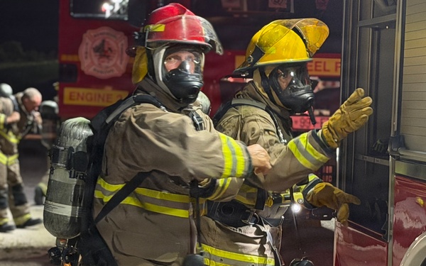A nighttime firefighting scene showing two fully equipped firefighters in the foreground wearing SCBA (Self-Contained Breathing Apparatus) gear, turnout suits, and full-face respirator masks. One firefighter wears a red helmet and the other a yellow helmet, both standing near a red fire engine. Additional firefighters are visible in the background near another fire apparatus.