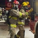 A nighttime firefighting scene showing two fully equipped firefighters in the foreground wearing SCBA (Self-Contained Breathing Apparatus) gear, turnout suits, and full-face respirator masks. One firefighter wears a red helmet and the other a yellow helmet, both standing near a red fire engine. Additional firefighters are visible in the background near another fire apparatus.