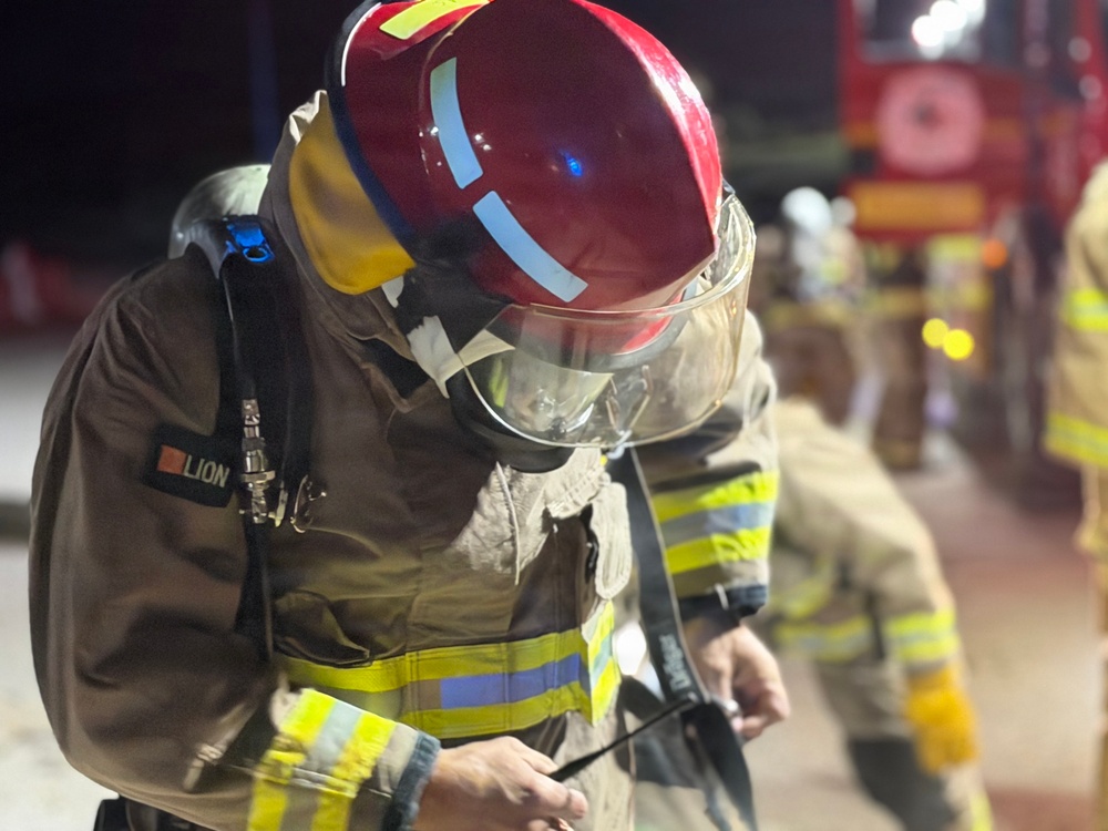 A firefighter in full protective gear is photographed at night, appearing to adjust or check equipment. The individual is wearing a red helmet with reflective strips, a face shield, SCBA (Self-Contained Breathing Apparatus) harness, and a tan/brown turnout suit with yellow and blue reflective trim branded 'LION'. A fire truck and at least one other firefighter are visible in the blurred background.