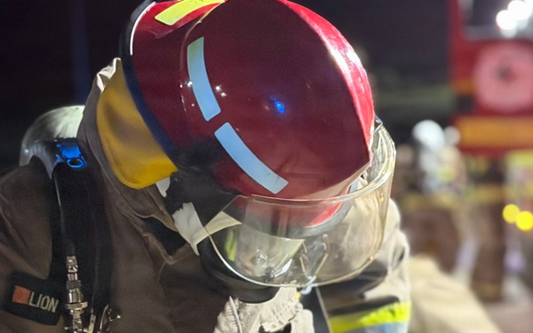 A firefighter in full protective gear is photographed at night, appearing to adjust or check equipment. The individual is wearing a red helmet with reflective strips, a face shield, SCBA (Self-Contained Breathing Apparatus) harness, and a tan/brown turnout suit with yellow and blue reflective trim branded 'LION'. A fire truck and at least one other firefighter are visible in the blurred background.
