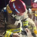 A firefighter in full protective gear is photographed at night, appearing to adjust or check equipment. The individual is wearing a red helmet with reflective strips, a face shield, SCBA (Self-Contained Breathing Apparatus) harness, and a tan/brown turnout suit with yellow and blue reflective trim branded 'LION'. A fire truck and at least one other firefighter are visible in the blurred background.