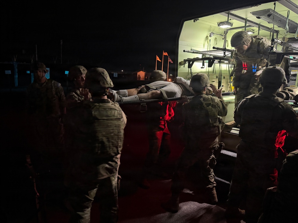 A nighttime military medical evacuation operation showing several soldiers in camouflage uniforms loading a person on a litter/stretcher into a military ambulance or medical vehicle. The scene is illuminated primarily by the vehicle's interior green lighting and red safety lights. This appears to be either a training exercise or an actual casualty evacuation (CASEVAC/MEDEVAC) operation.