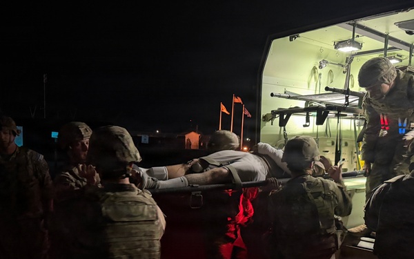 A nighttime military medical evacuation operation showing several soldiers in camouflage uniforms loading a person on a litter/stretcher into a military ambulance or medical vehicle. The scene is illuminated primarily by the vehicle's interior green lighting and red safety lights. This appears to be either a training exercise or an actual casualty evacuation (CASEVAC/MEDEVAC) operation.