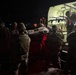 A nighttime military medical evacuation operation showing several soldiers in camouflage uniforms loading a person on a litter/stretcher into a military ambulance or medical vehicle. The scene is illuminated primarily by the vehicle's interior green lighting and red safety lights. This appears to be either a training exercise or an actual casualty evacuation (CASEVAC/MEDEVAC) operation.