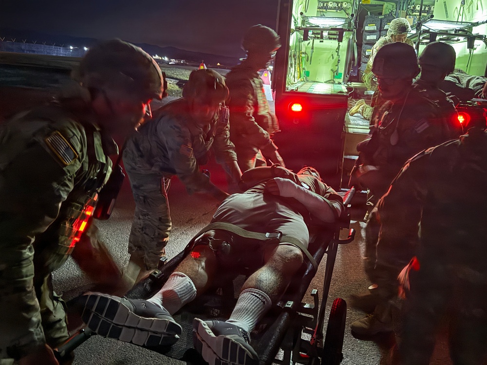 A nighttime military medical evacuation operation showing uniformed personnel transferring a patient on a stretcher near a vehicle with red taillights. Multiple soldiers wearing combat helmets and camouflage uniforms are coordinating the movement of the casualty. The scene appears to take place on a tarmac or paved surface, likely at a military airfield or forward operating base.