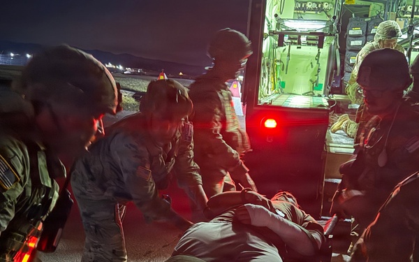 A nighttime military medical evacuation operation showing uniformed personnel transferring a patient on a stretcher near a vehicle with red taillights. Multiple soldiers wearing combat helmets and camouflage uniforms are coordinating the movement of the casualty. The scene appears to take place on a tarmac or paved surface, likely at a military airfield or forward operating base.