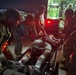 A nighttime military medical evacuation operation showing uniformed personnel transferring a patient on a stretcher near a vehicle with red taillights. Multiple soldiers wearing combat helmets and camouflage uniforms are coordinating the movement of the casualty. The scene appears to take place on a tarmac or paved surface, likely at a military airfield or forward operating base.