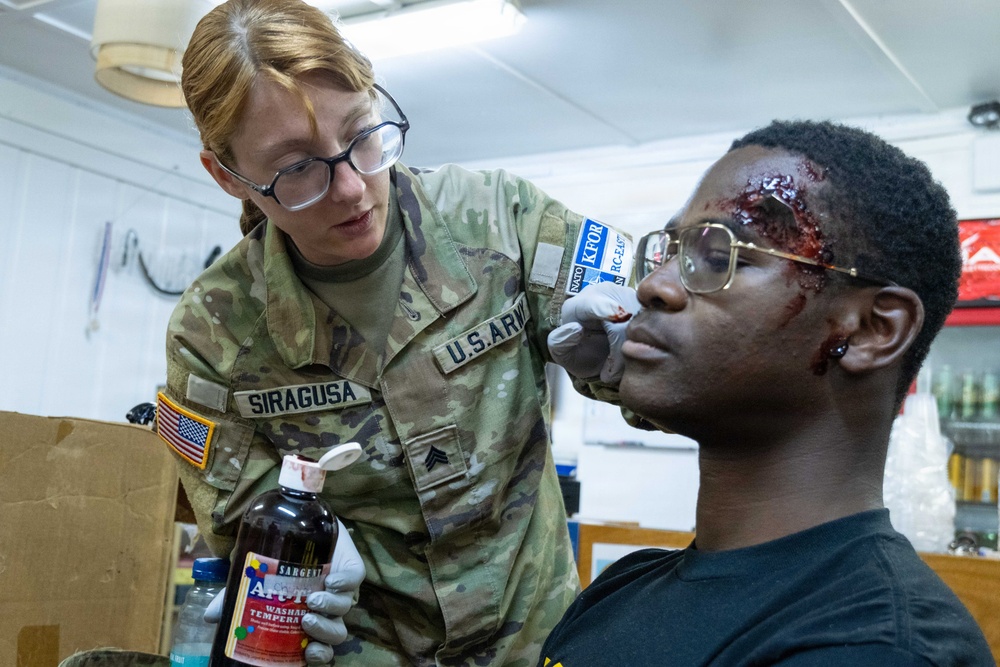 A U.S. Army soldier named Siragusa, wearing a KFOR (Kosovo Force) patch, applies simulated wound makeup to a seated individual using red tempera paint. The 'wounds' on the subject's forehead and face are clearly artificial, created for a training exercise or medical simulation scenario. The soldier is wearing latex gloves and holding a bottle of Sargent Art washable tempera paint.