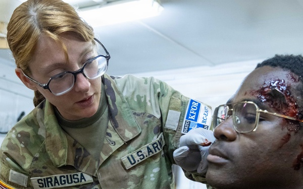 A U.S. Army soldier named Siragusa, wearing a KFOR (Kosovo Force) patch, applies simulated wound makeup to a seated individual using red tempera paint. The 'wounds' on the subject's forehead and face are clearly artificial, created for a training exercise or medical simulation scenario. The soldier is wearing latex gloves and holding a bottle of Sargent Art washable tempera paint.