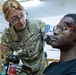 A U.S. Army soldier named Siragusa, wearing a KFOR (Kosovo Force) patch, applies simulated wound makeup to a seated individual using red tempera paint. The 'wounds' on the subject's forehead and face are clearly artificial, created for a training exercise or medical simulation scenario. The soldier is wearing latex gloves and holding a bottle of Sargent Art washable tempera paint.