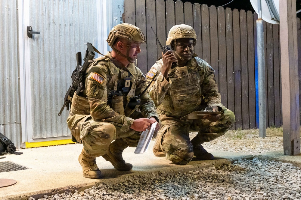 Two U.S. Army soldiers in MultiCam (OCP) camouflage uniforms are crouching near a building structure during what appears to be a nighttime training exercise or operation. The soldier on the right, whose name tape reads 'LALOR', is speaking into a handheld radio while holding a document or map. The soldier on the left is also reviewing a paper document and is equipped with a rifle and additional gear.