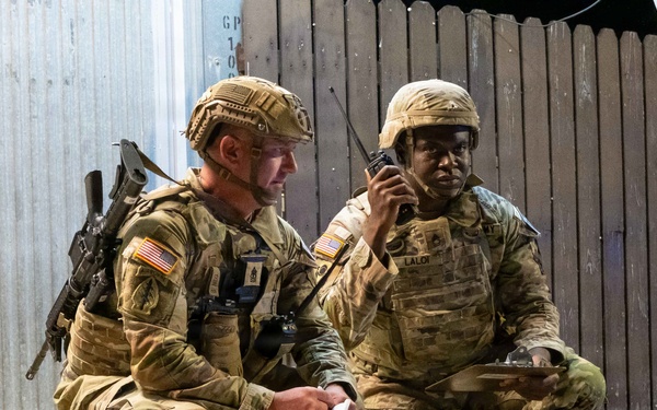 Two U.S. Army soldiers in MultiCam (OCP) camouflage uniforms are crouching near a building structure during what appears to be a nighttime training exercise or operation. The soldier on the right, whose name tape reads 'LALOR', is speaking into a handheld radio while holding a document or map. The soldier on the left is also reviewing a paper document and is equipped with a rifle and additional gear.