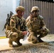 Two U.S. Army soldiers in MultiCam (OCP) camouflage uniforms are crouching near a building structure during what appears to be a nighttime training exercise or operation. The soldier on the right, whose name tape reads 'LALOR', is speaking into a handheld radio while holding a document or map. The soldier on the left is also reviewing a paper document and is equipped with a rifle and additional gear.