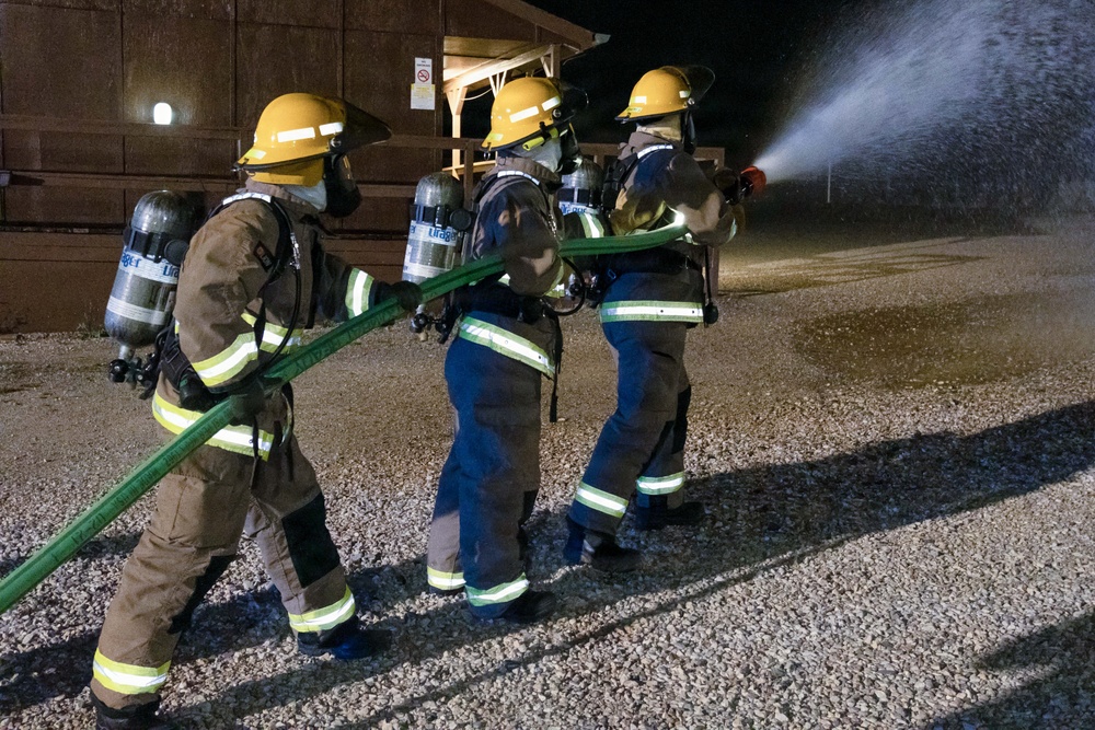 Three firefighters in full protective gear are conducting a nighttime hose training exercise on a gravel surface. They are working together to control a green fire hose that is actively discharging a high-pressure water stream. All personnel are equipped with SCBA (Self-Contained Breathing Apparatus) units and yellow helmets, suggesting this is a structured training drill.