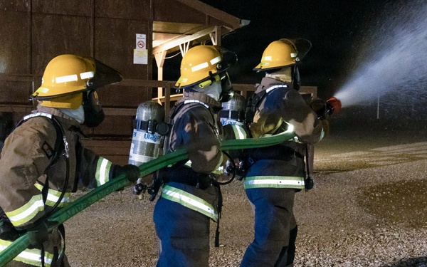 Three firefighters in full protective gear are conducting a nighttime hose training exercise on a gravel surface. They are working together to control a green fire hose that is actively discharging a high-pressure water stream. All personnel are equipped with SCBA (Self-Contained Breathing Apparatus) units and yellow helmets, suggesting this is a structured training drill.