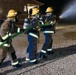 Three firefighters in full protective gear are conducting a nighttime hose training exercise on a gravel surface. They are working together to control a green fire hose that is actively discharging a high-pressure water stream. All personnel are equipped with SCBA (Self-Contained Breathing Apparatus) units and yellow helmets, suggesting this is a structured training drill.